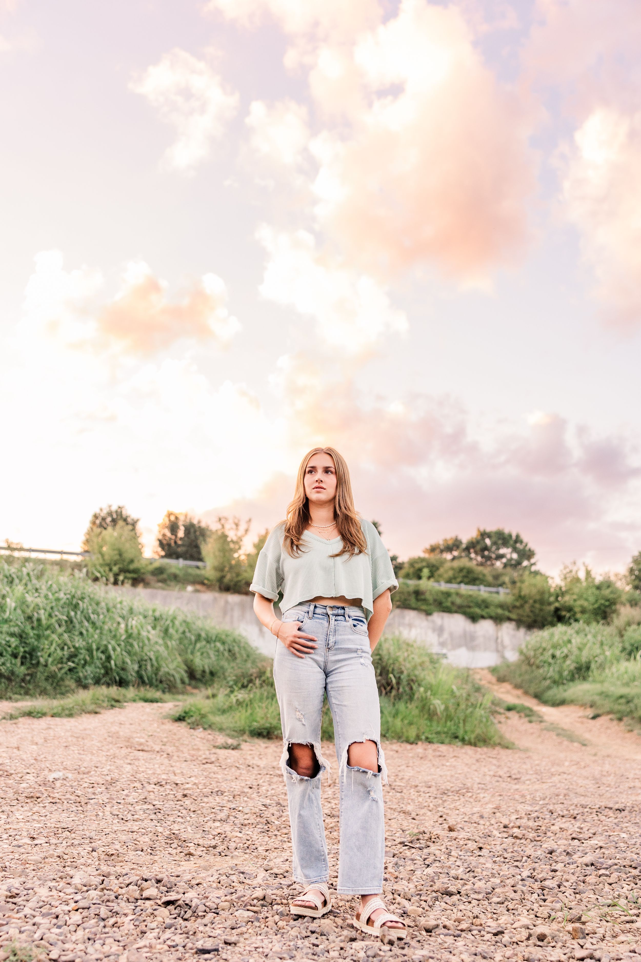 Woman in distressed jeans poses against sunset sky on dirt path near greenery.
