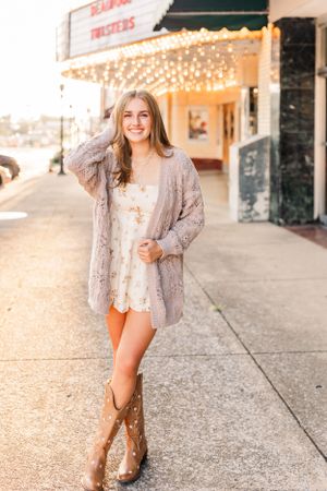 Woman in cowboy boots and cardigan posing on sidewalk near storefront with string lights.