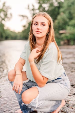 Woman in casual attire crouching on rocky riverbank during golden hour.