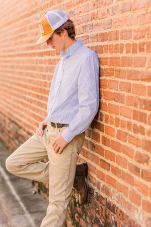Young man in baseball cap and button-down shirt leaning against brick wall.