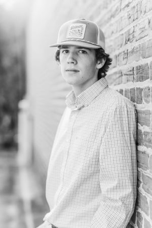 Black and white portrait of young man wearing trucker hat against brick wall.