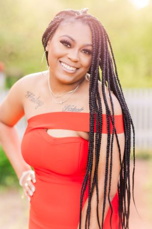 Woman with long braided hair wearing red dress smiles brightly in outdoor setting.