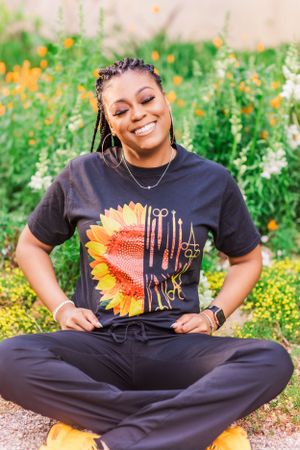 Casual outdoor portrait showing someone in a sunflower graphic t-shirt sitting cross-legged among flowers.