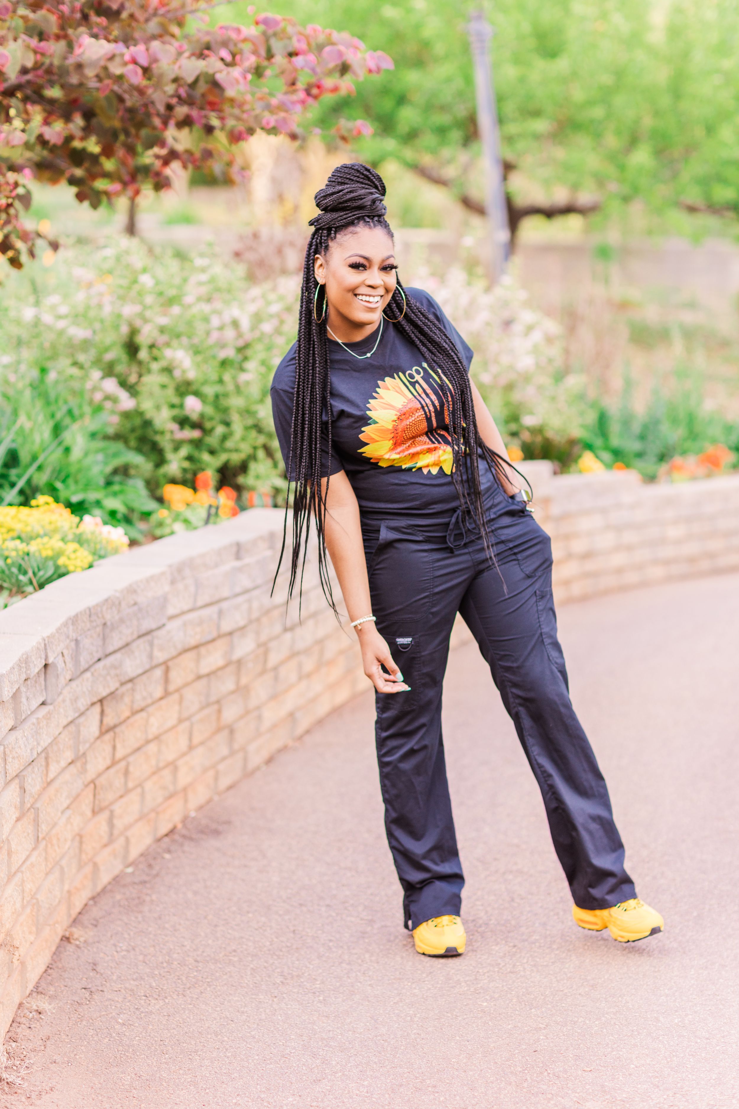 Stylish outdoor pose featuring someone in a sunflower t-shirt and black pants against a garden backdrop.