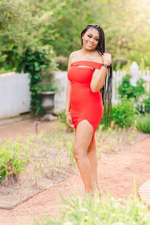 Woman in vibrant red dress poses outdoors in garden setting with natural sunlight.