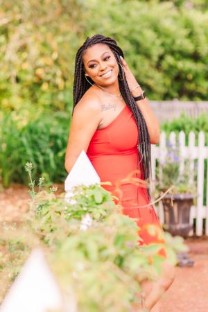 Woman in red dress poses elegantly among garden flowers with long braided hairstyle.
