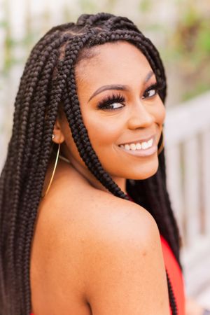 Portrait of someone with long box braids and a bright smile wearing a red top against a blurred background.