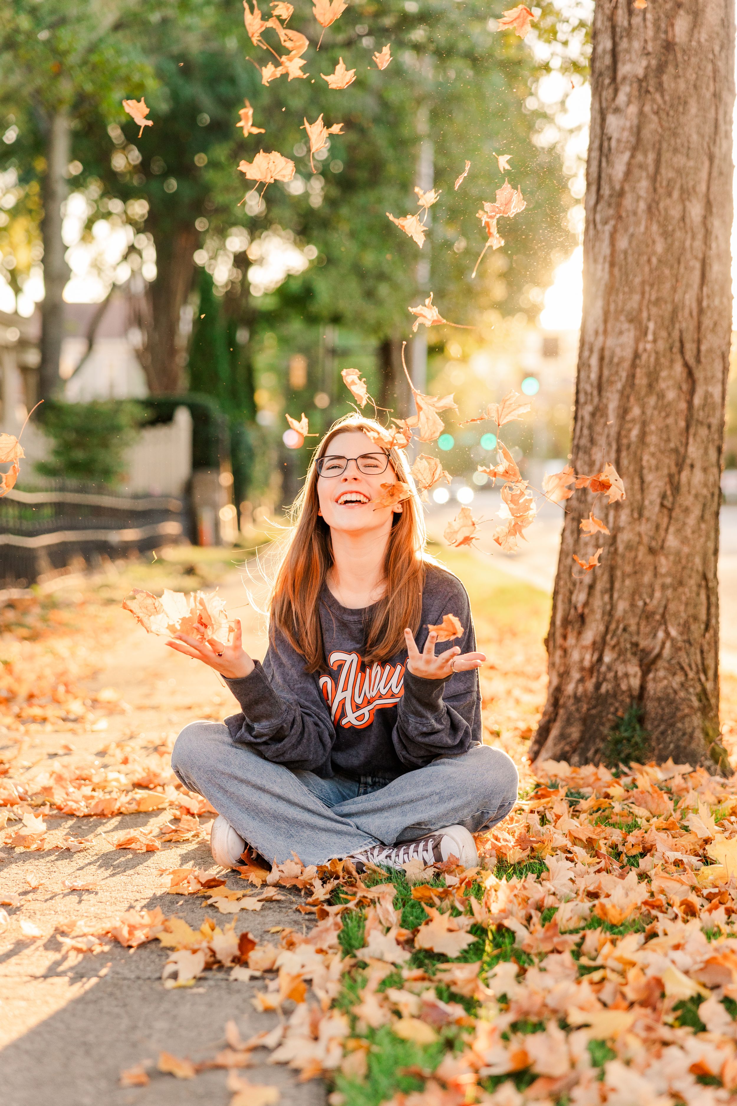 Person sitting cross-legged on fallen autumn leaves throwing foliage in the air during golden hour.