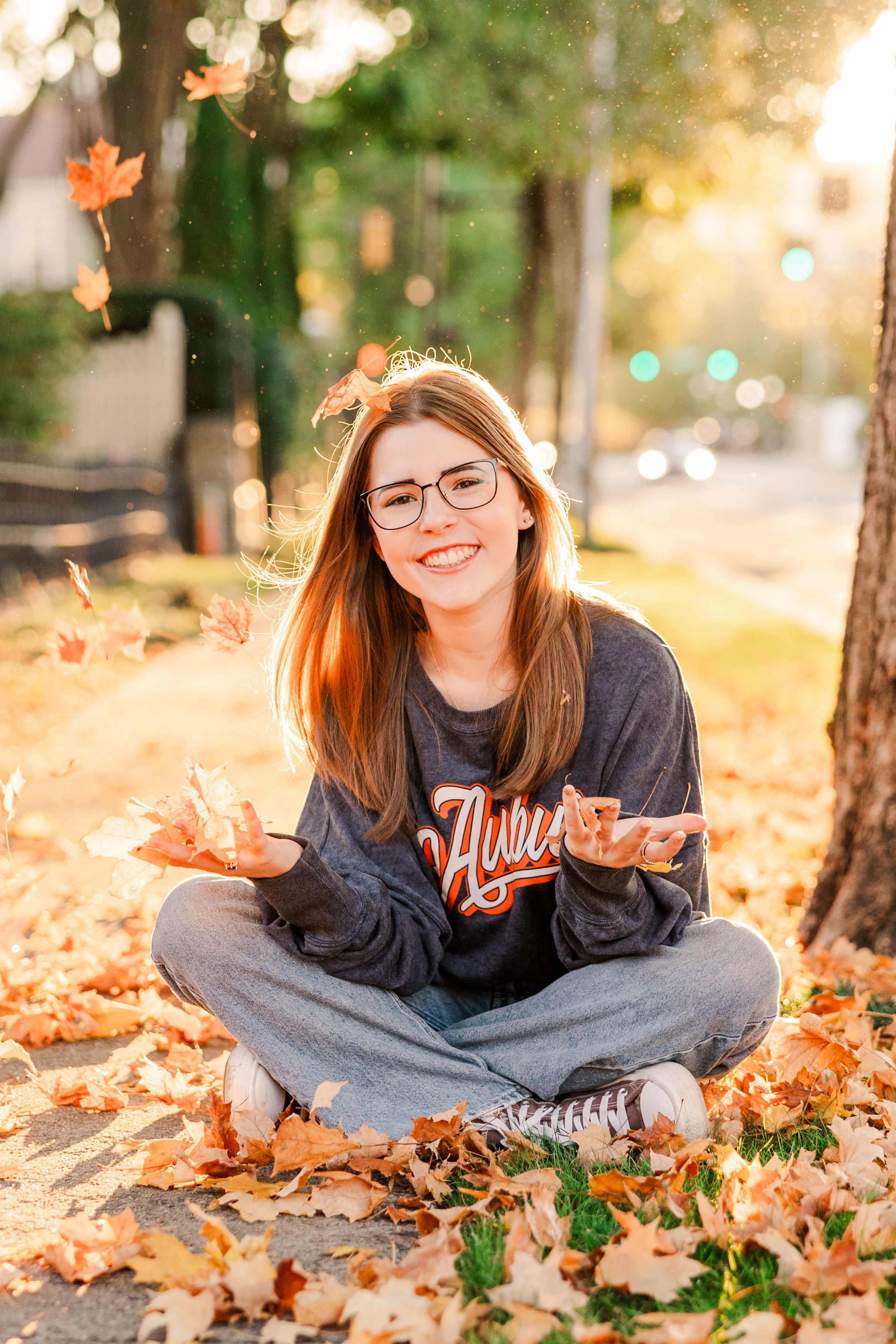 Person in navy sweatshirt sitting on ground surrounded by falling autumn leaves at sunset.
