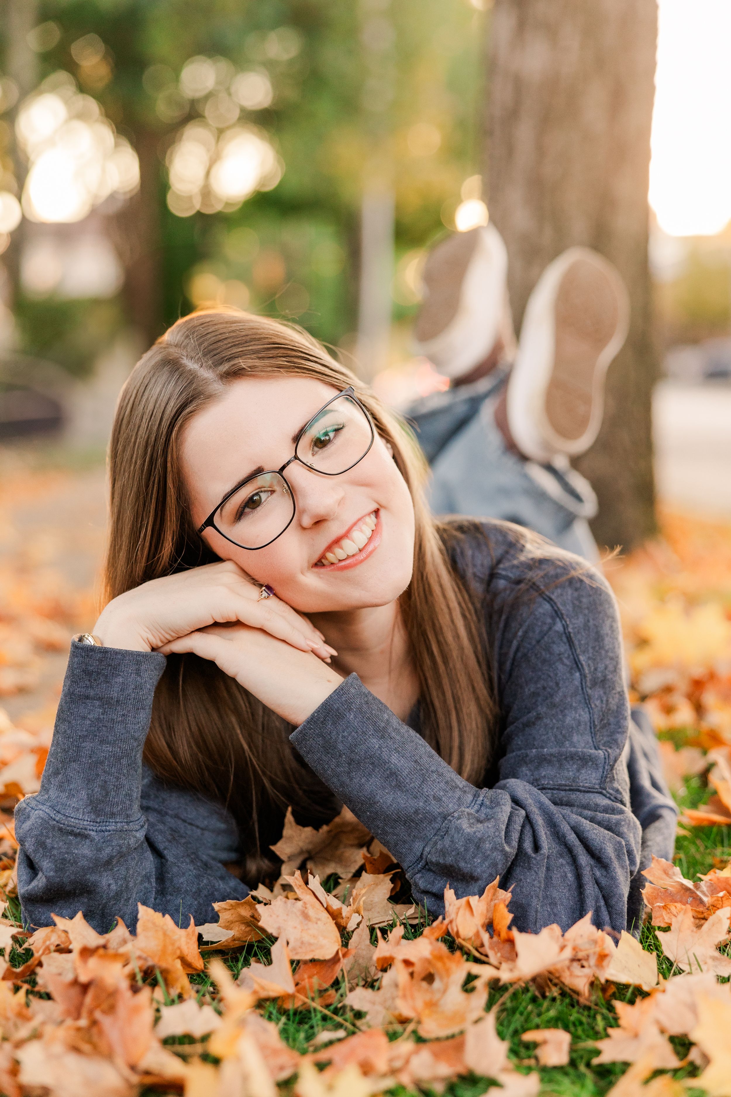 Person lying in autumn leaves wearing gray sweater and glasses with joyful expression.