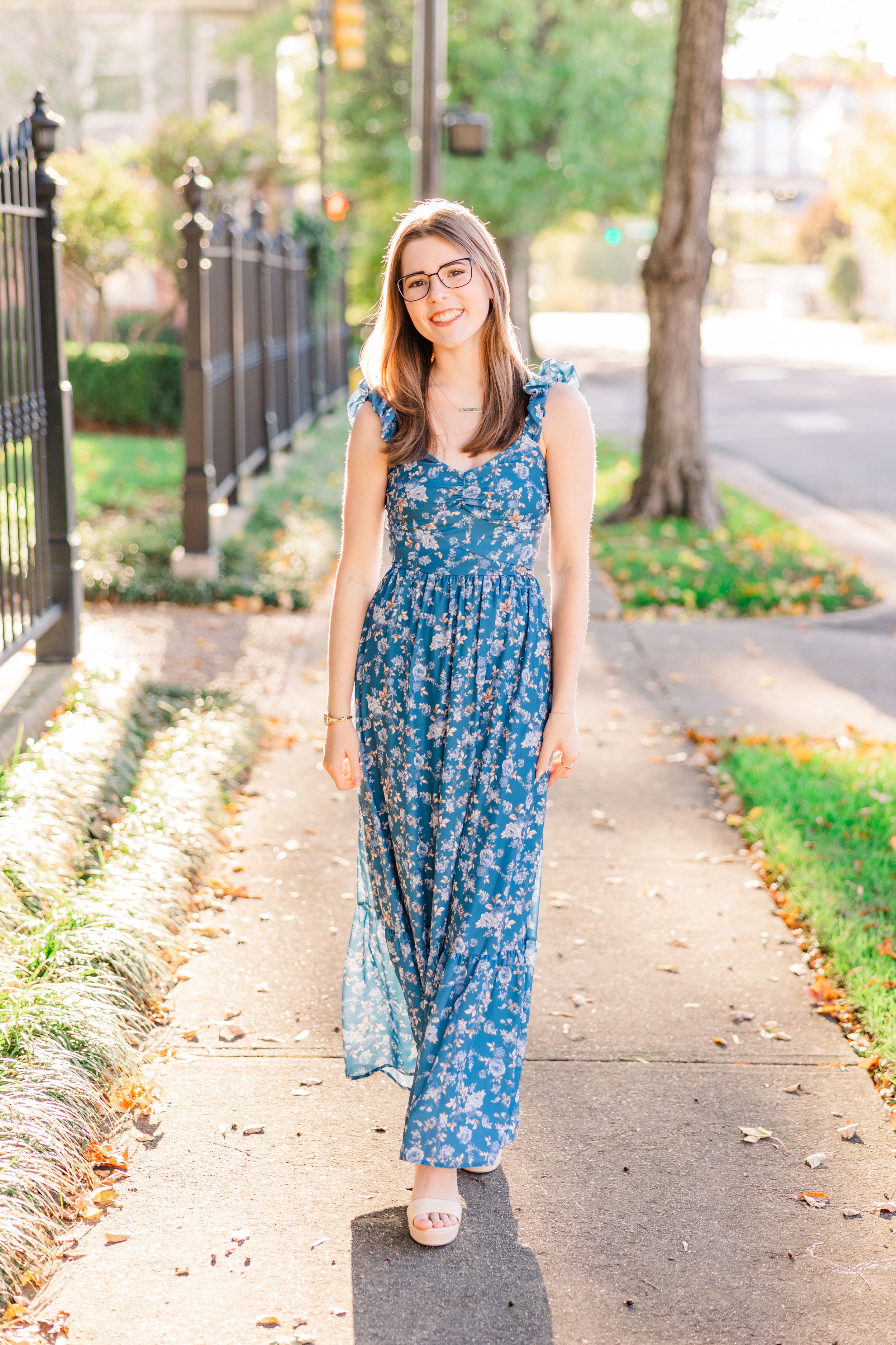 Woman in floral blue maxi dress smiling on sunny sidewalk.