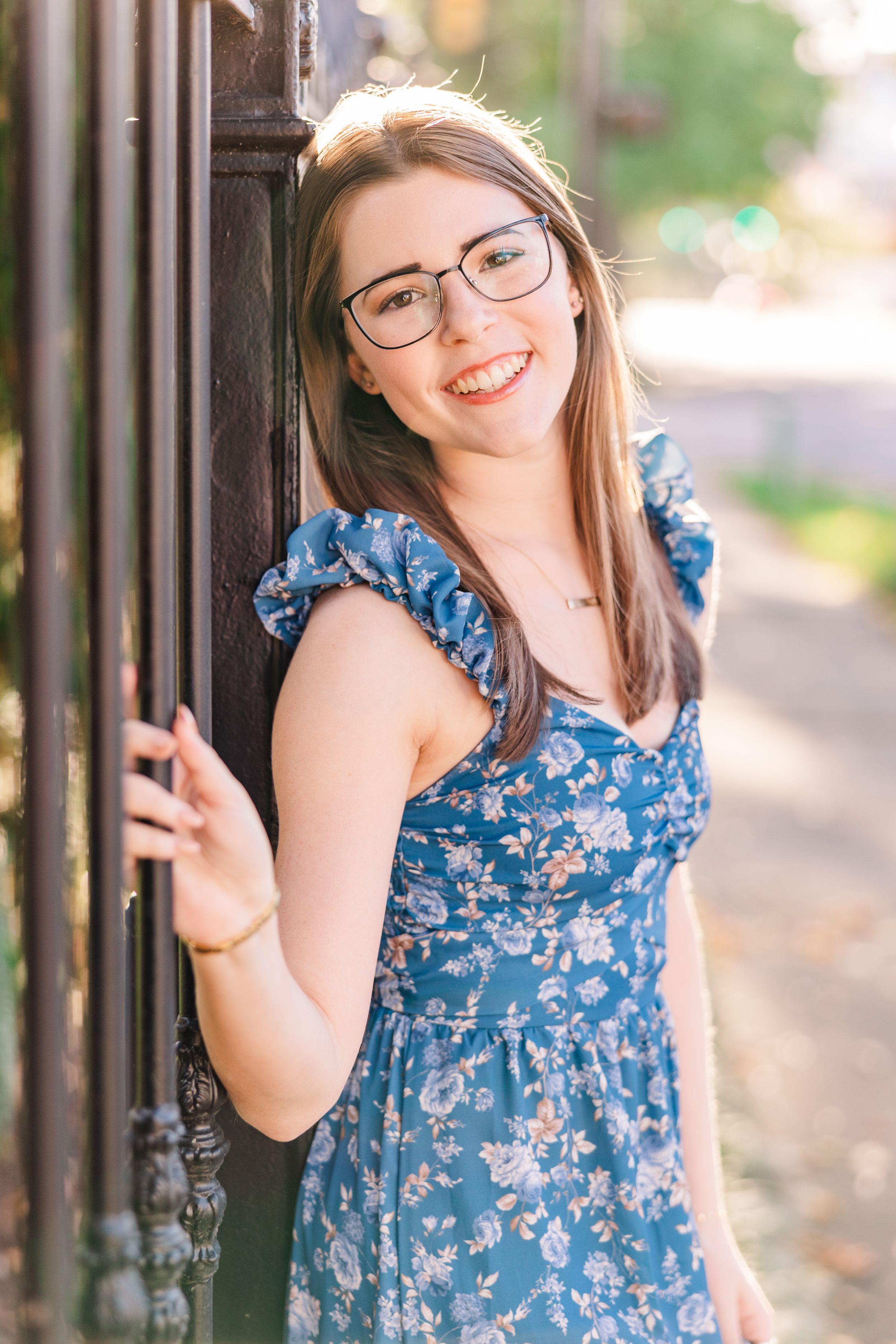 Young woman in blue floral dress leaning against iron fence post.