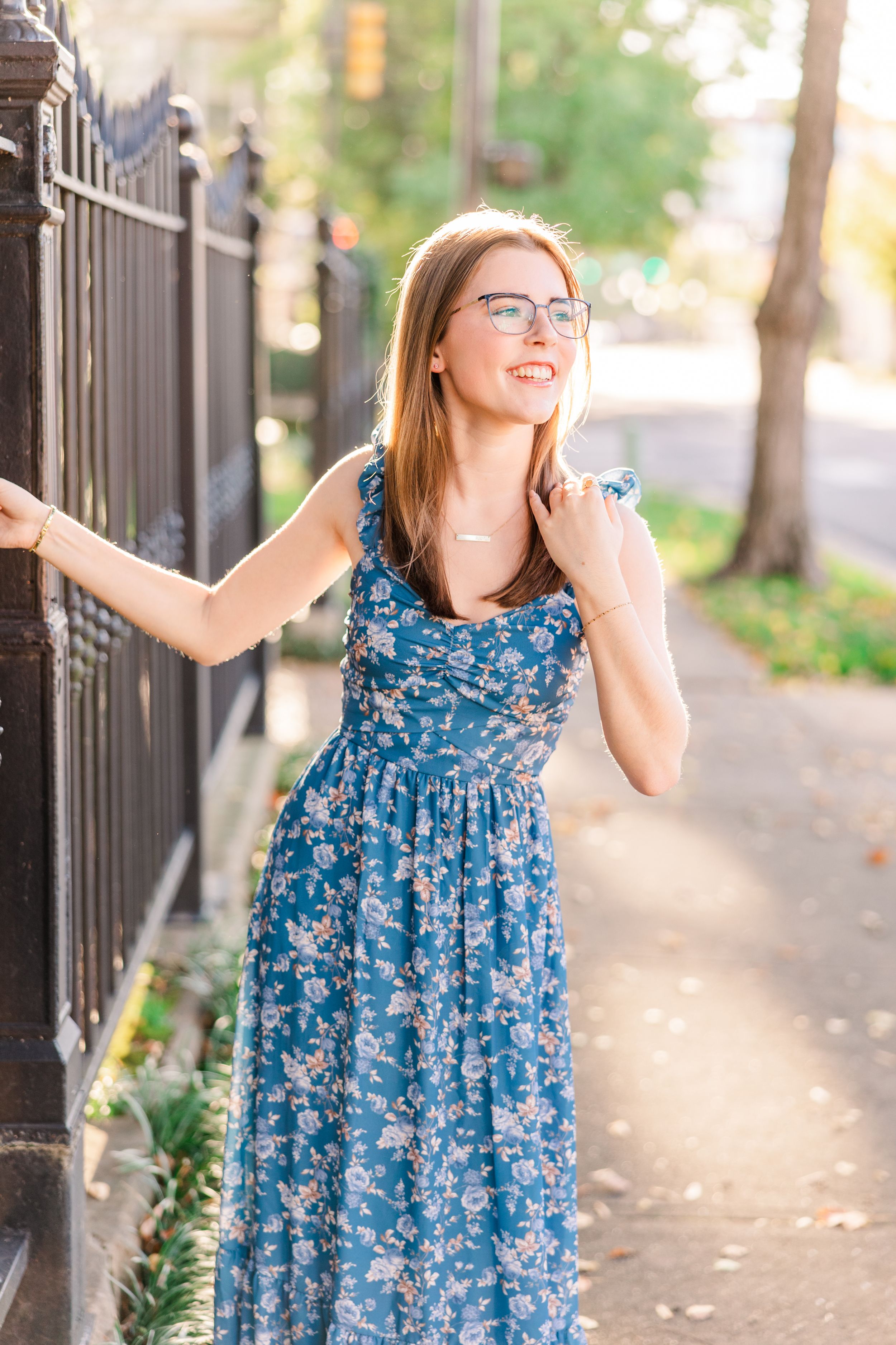 Woman in glasses wearing blue floral maxi dress posing by fence.