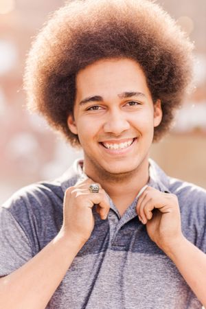 Young person with natural afro hairstyle wearing a grey polo shirt smiles warmly at the camera.