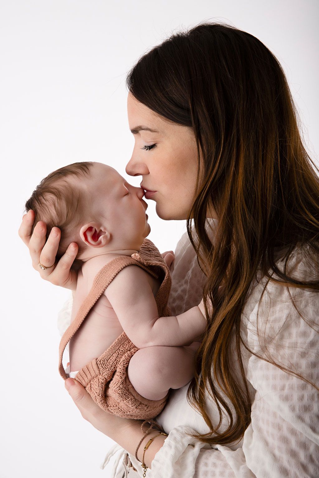 Intimate portrait of parent cradling and kissing newborn baby wearing brown in soft lighting.