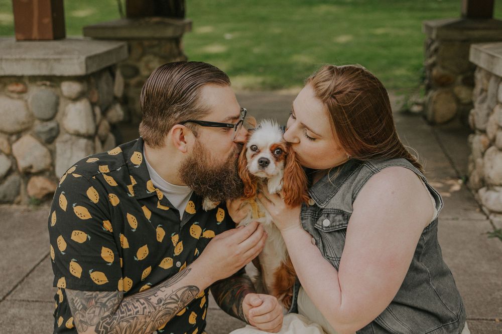 A couple shares affectionate moments with their Cavalier King Charles Spaniel in an outdoor setting.