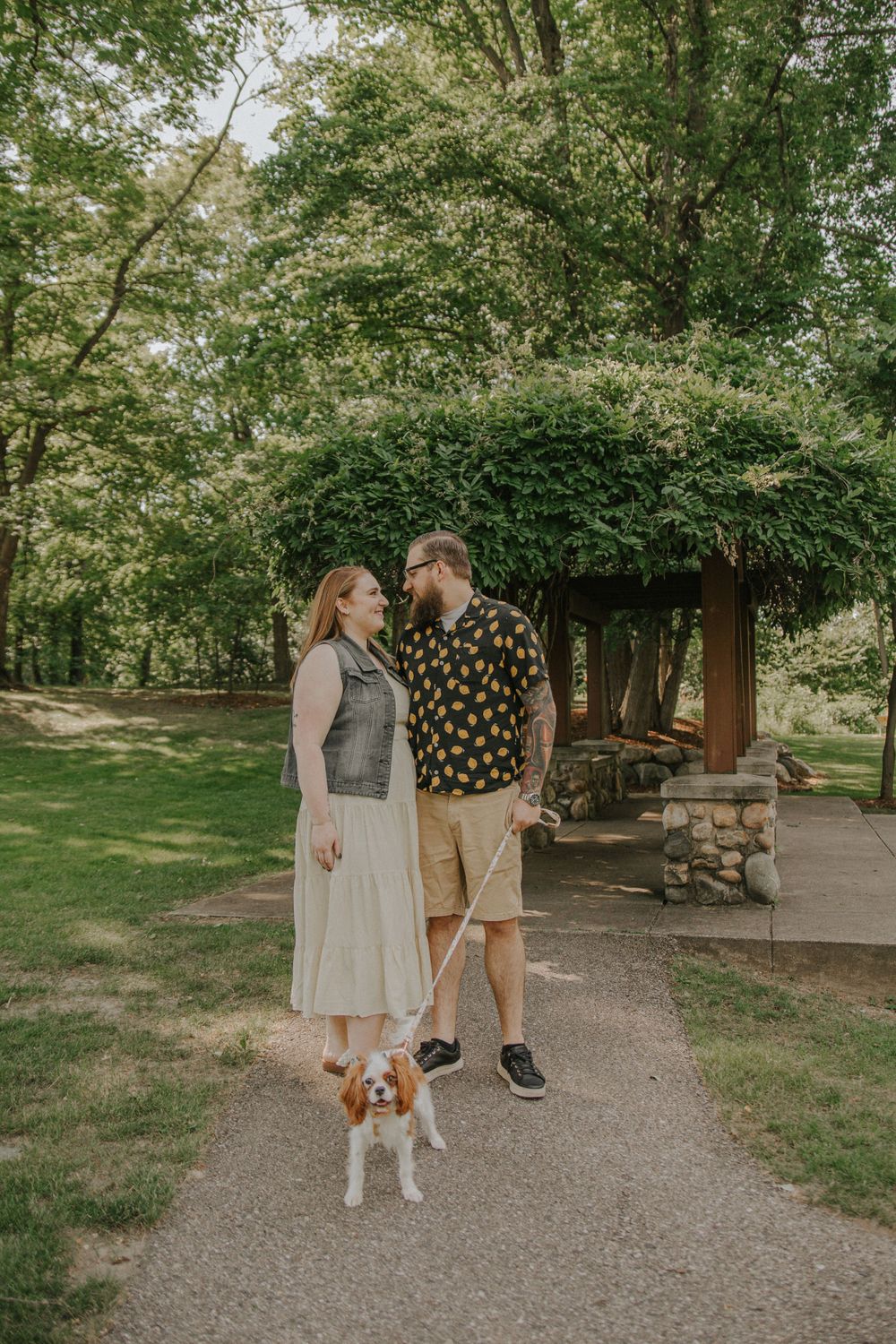 A couple shares a tender moment on a tree-lined path with their small dog.