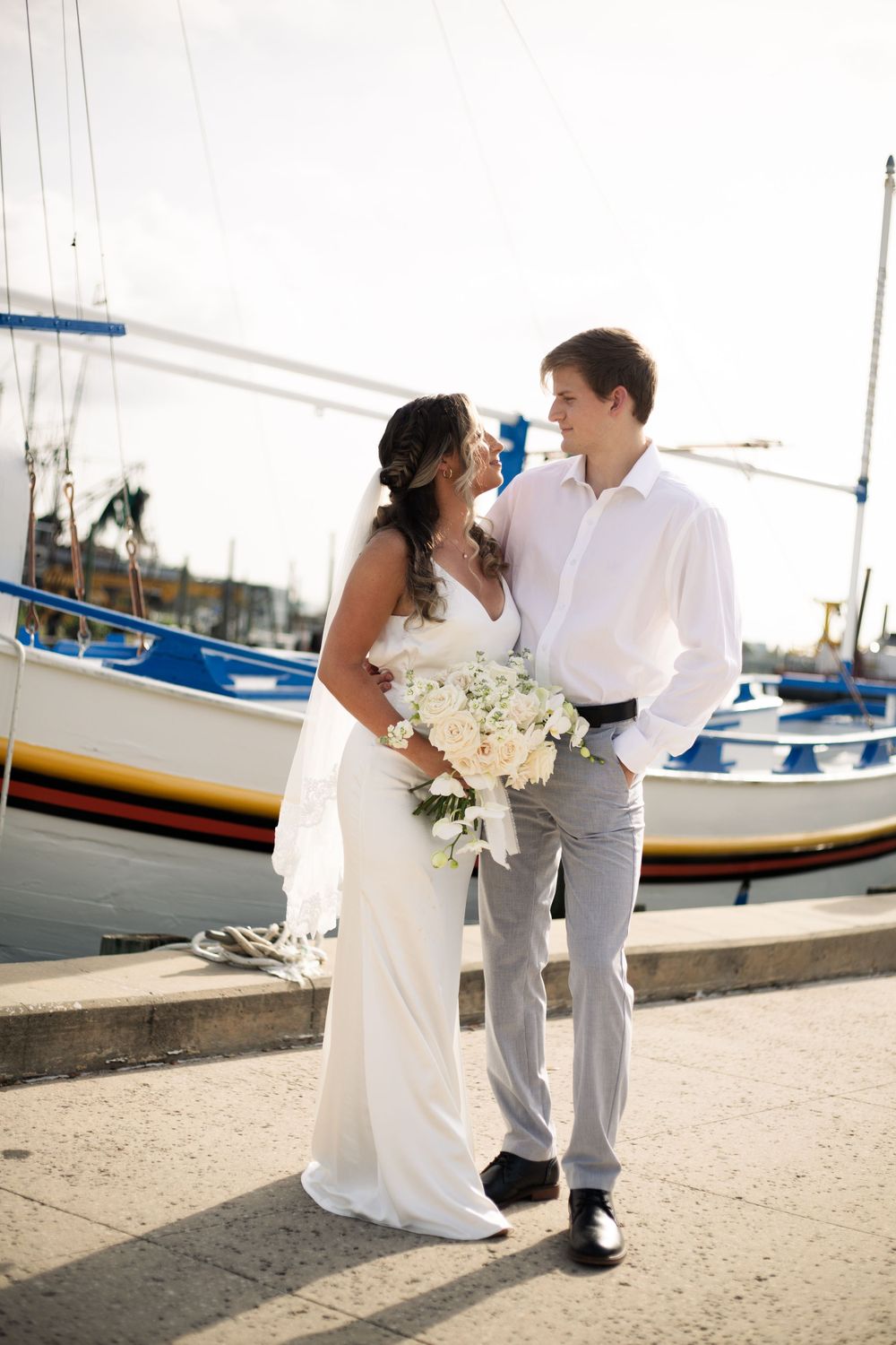 A newlywed couple poses by sailboats at a marina, holding an elegant white bouquet.