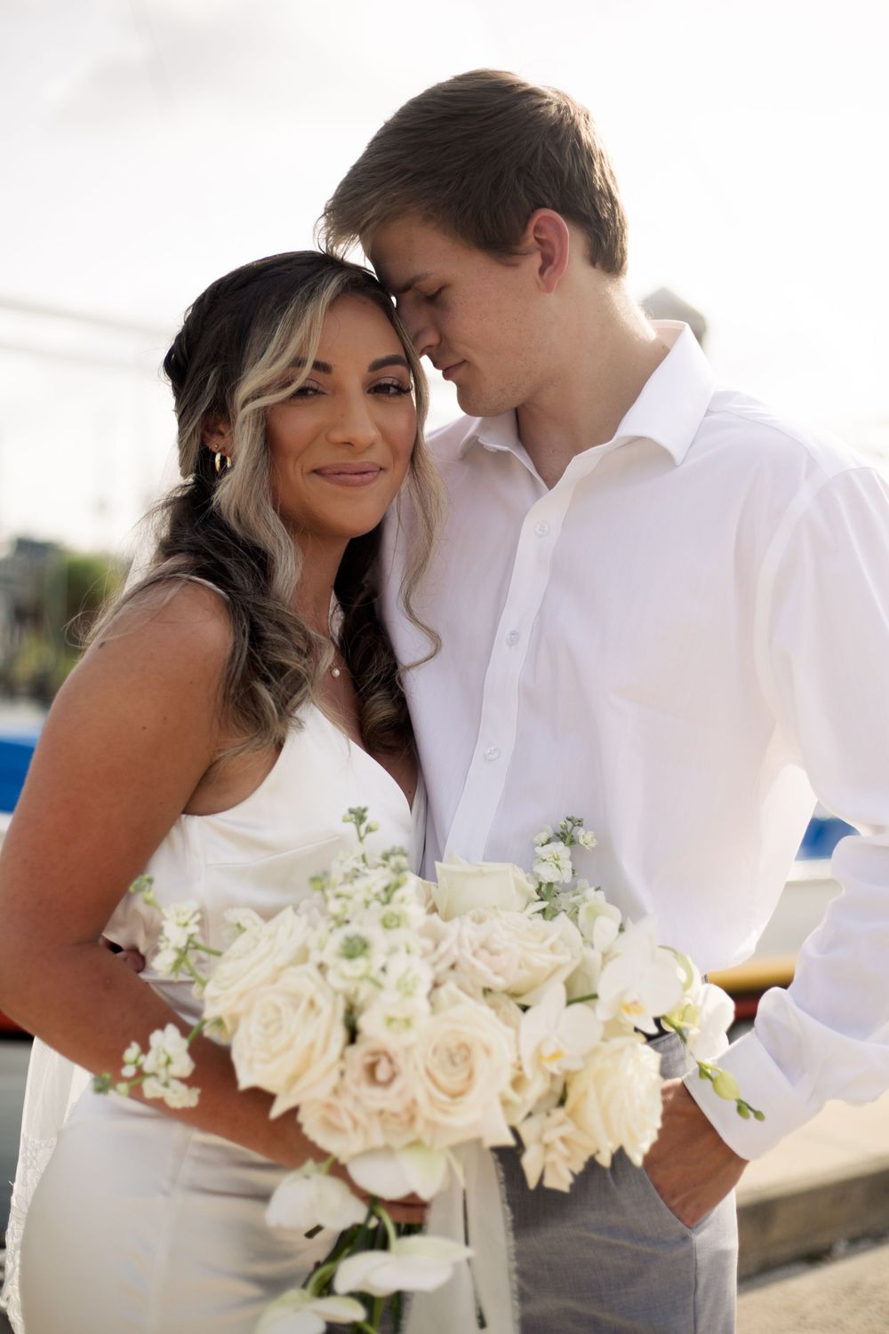 A close-up portrait of a couple sharing an intimate moment with white roses at a marina.