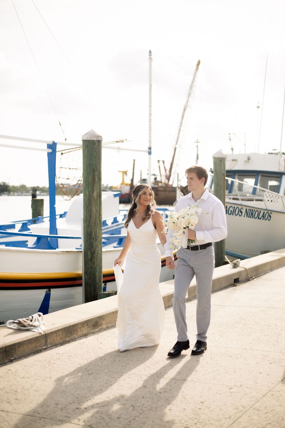 A couple strolls along a marina boardwalk holding a bouquet of white flowers.