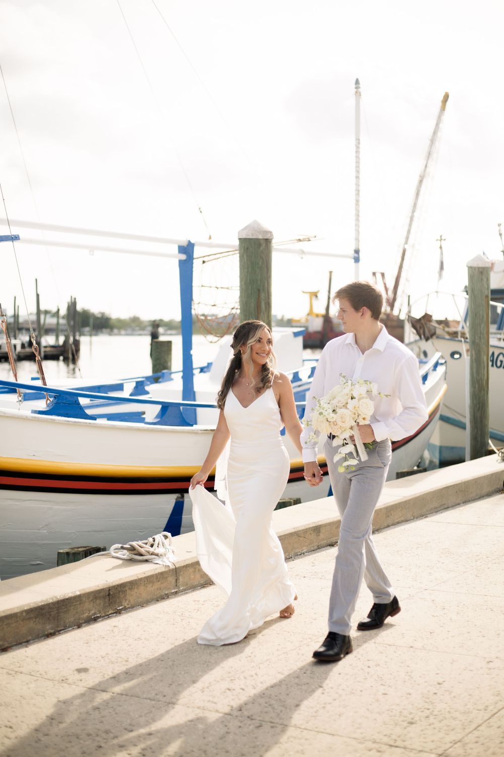 A couple takes a romantic walk along a sun-lit marina dock with sailboats in view.