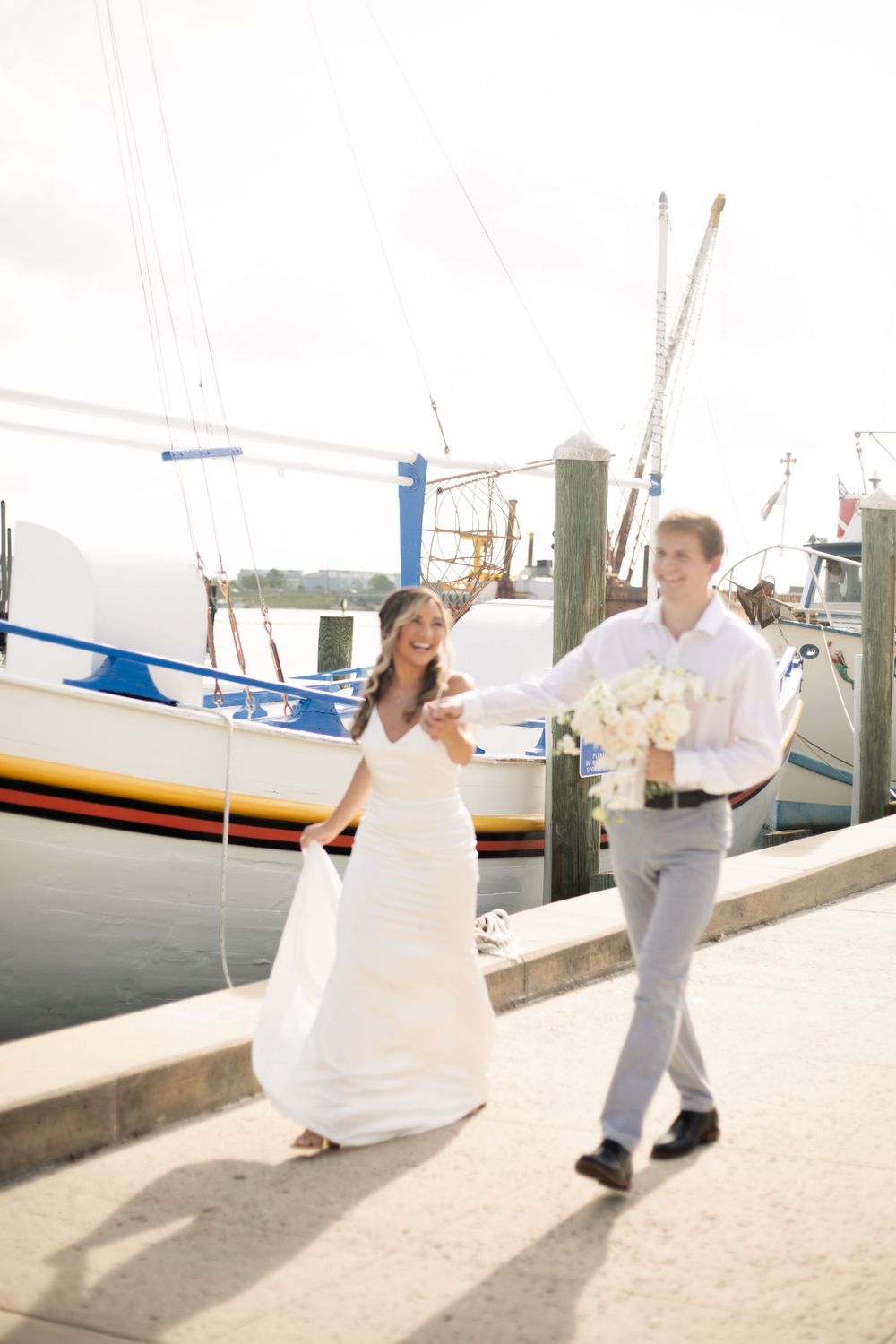 A couple in wedding attire walks joyfully along a marina dock with boats in the background.