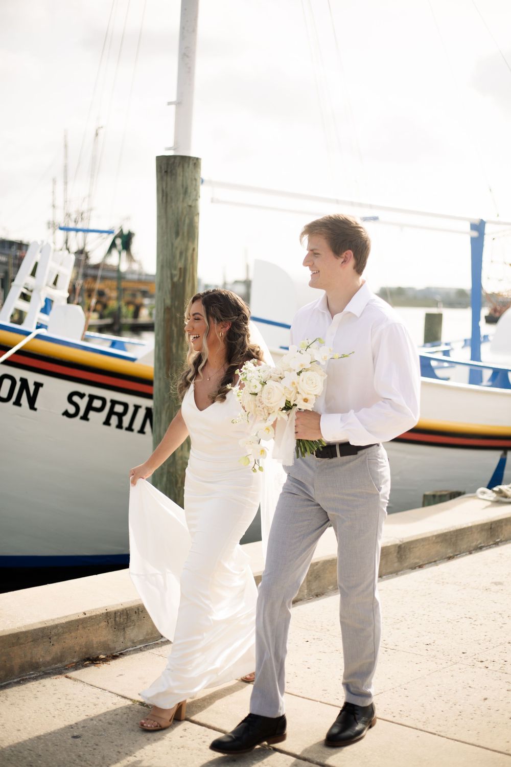 Newlyweds share a playful moment while walking near boats at a waterfront marina.