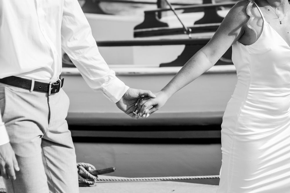 Black and white photo of newlyweds holding hands by a boat.