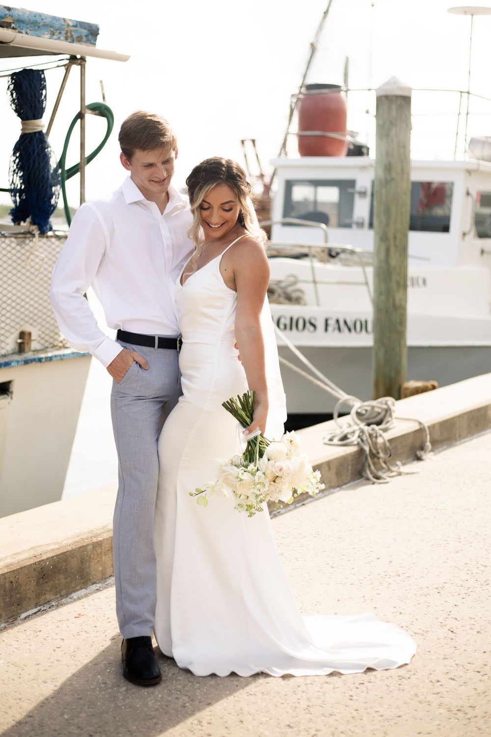 Newlyweds pose on a dock next to a boat wearing elegant white wedding attire and holding a bouquet of flowers.