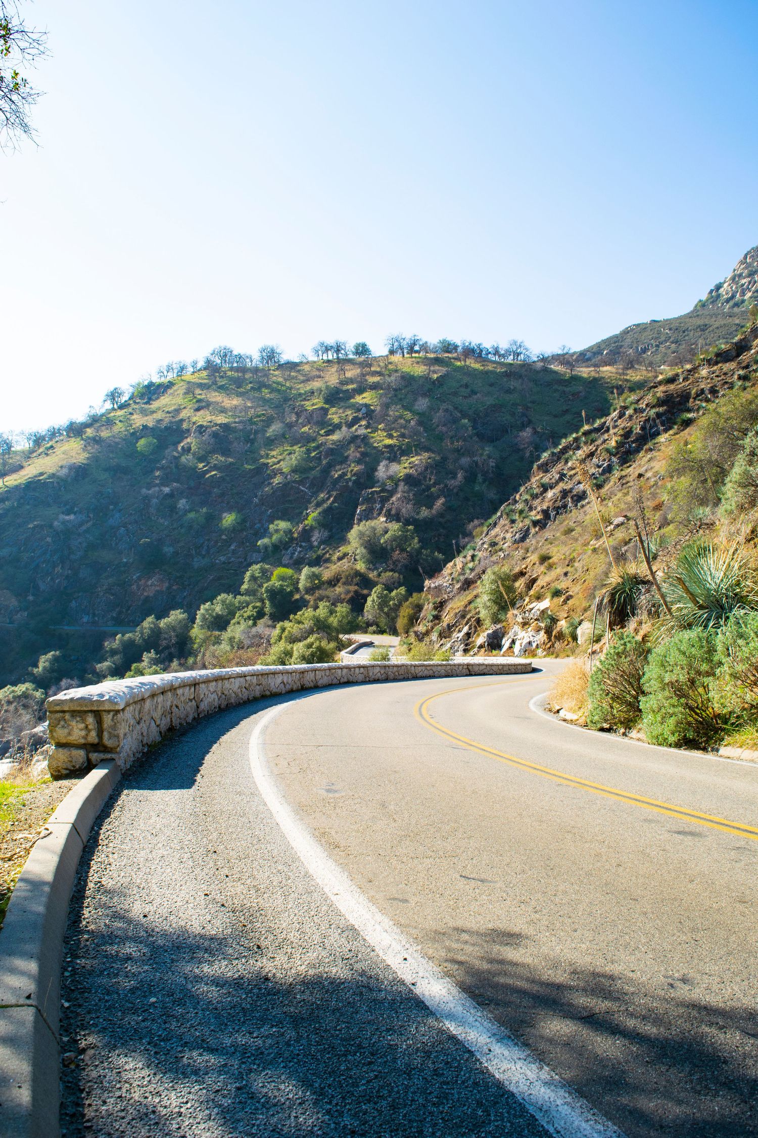 Winding mountain road with stone barrier curves along steep hillside under bright blue sky.