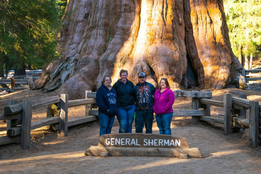 Visitors pose in front of the massive General Sherman sequoia tree with a wooden fence and sign in Sequoia National Park.