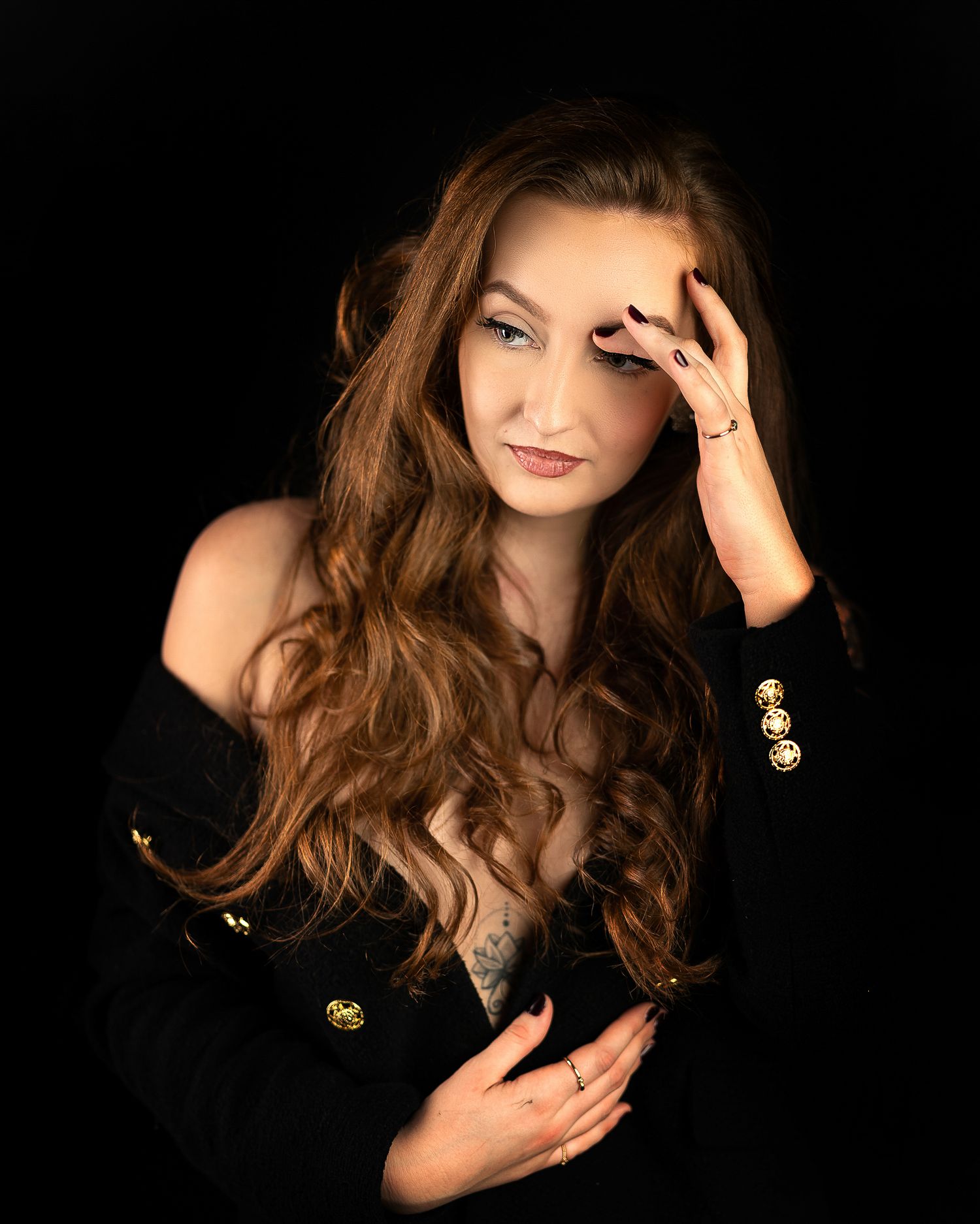 Woman with long auburn wavy hair posing in black off-shoulder jacket against dark background.