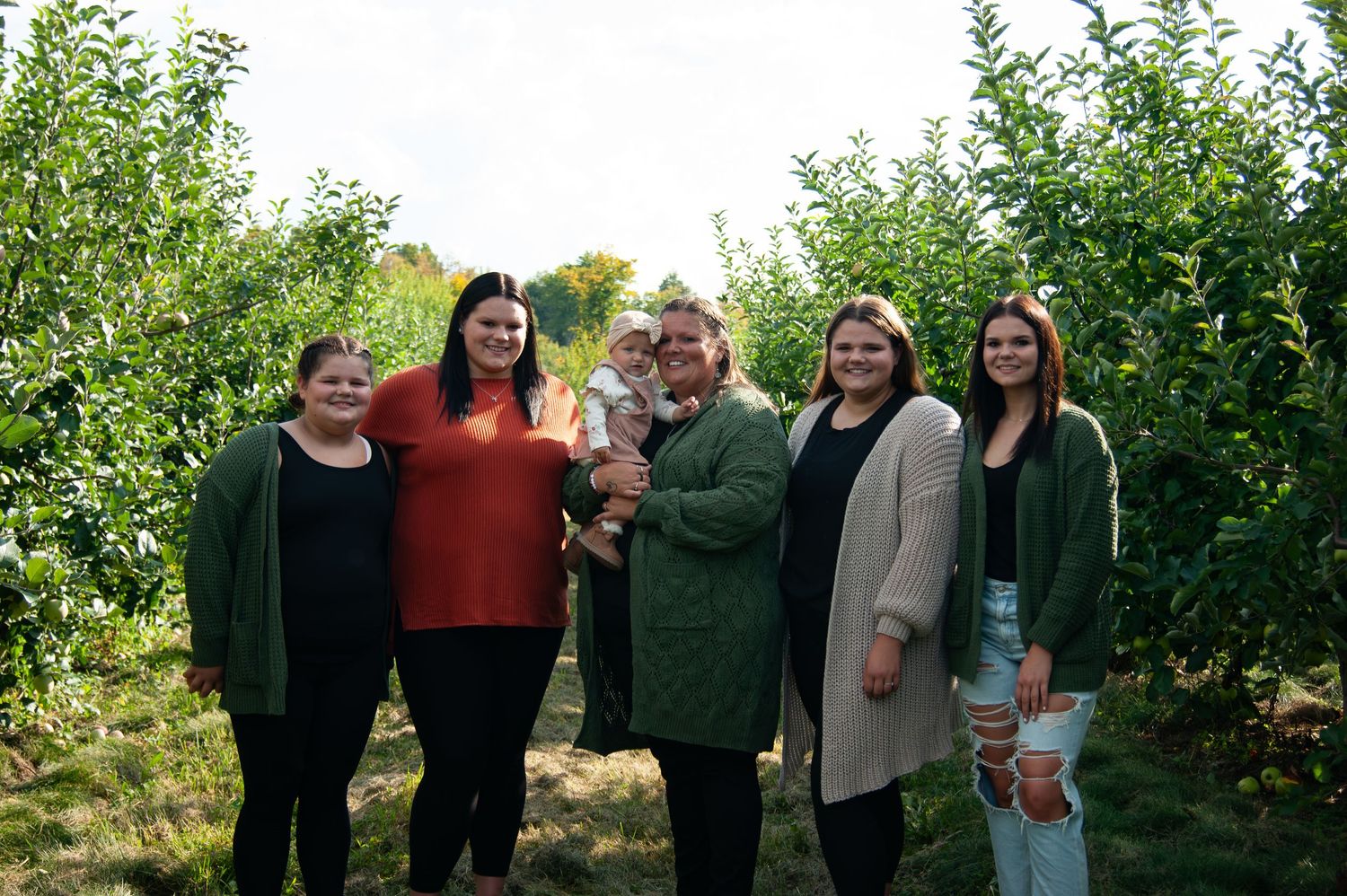 A group gathers together for a casual photo in an apple orchard on a sunny day.
