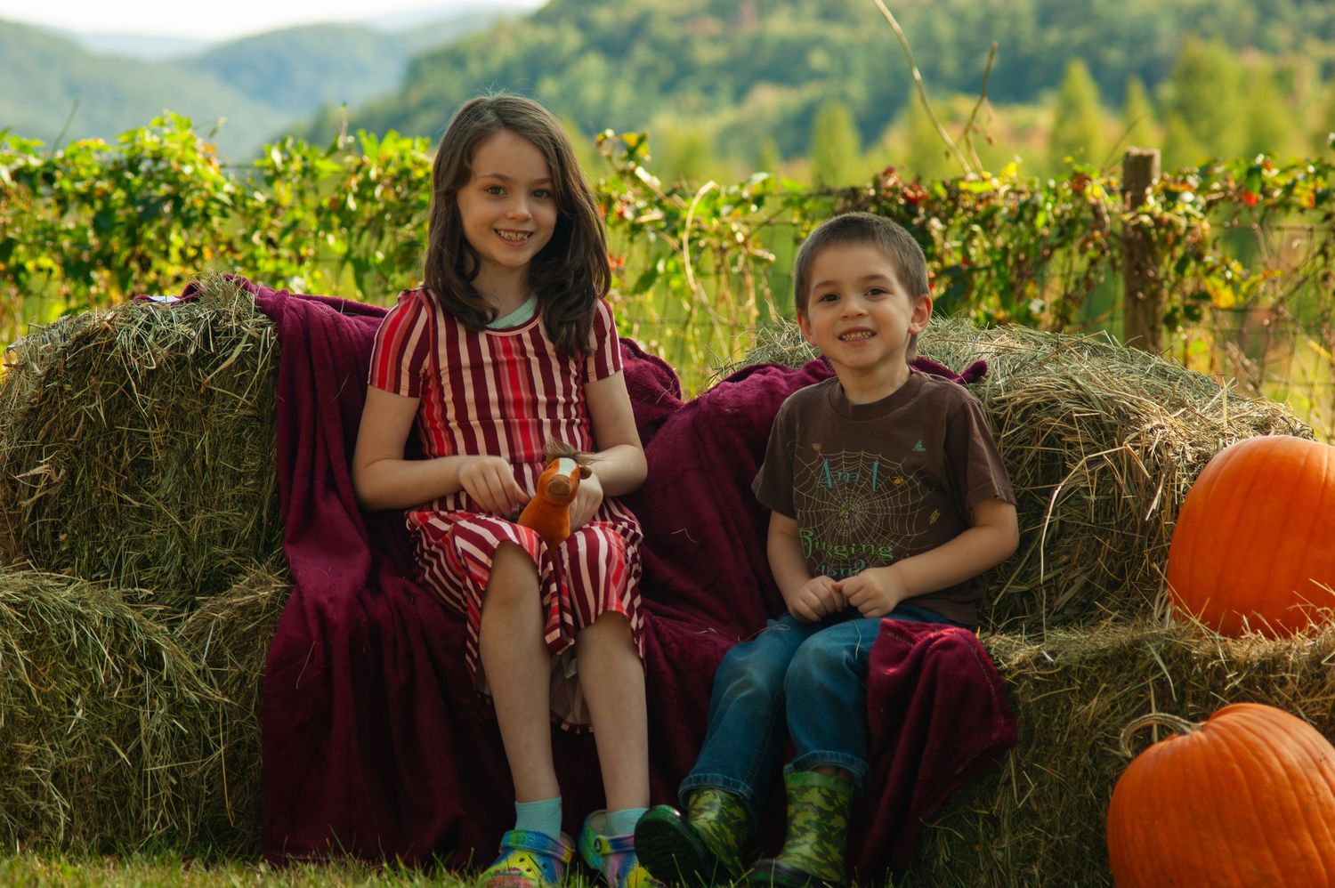 Two people sit on hay bales with orange pumpkins in a scenic mountain setting.