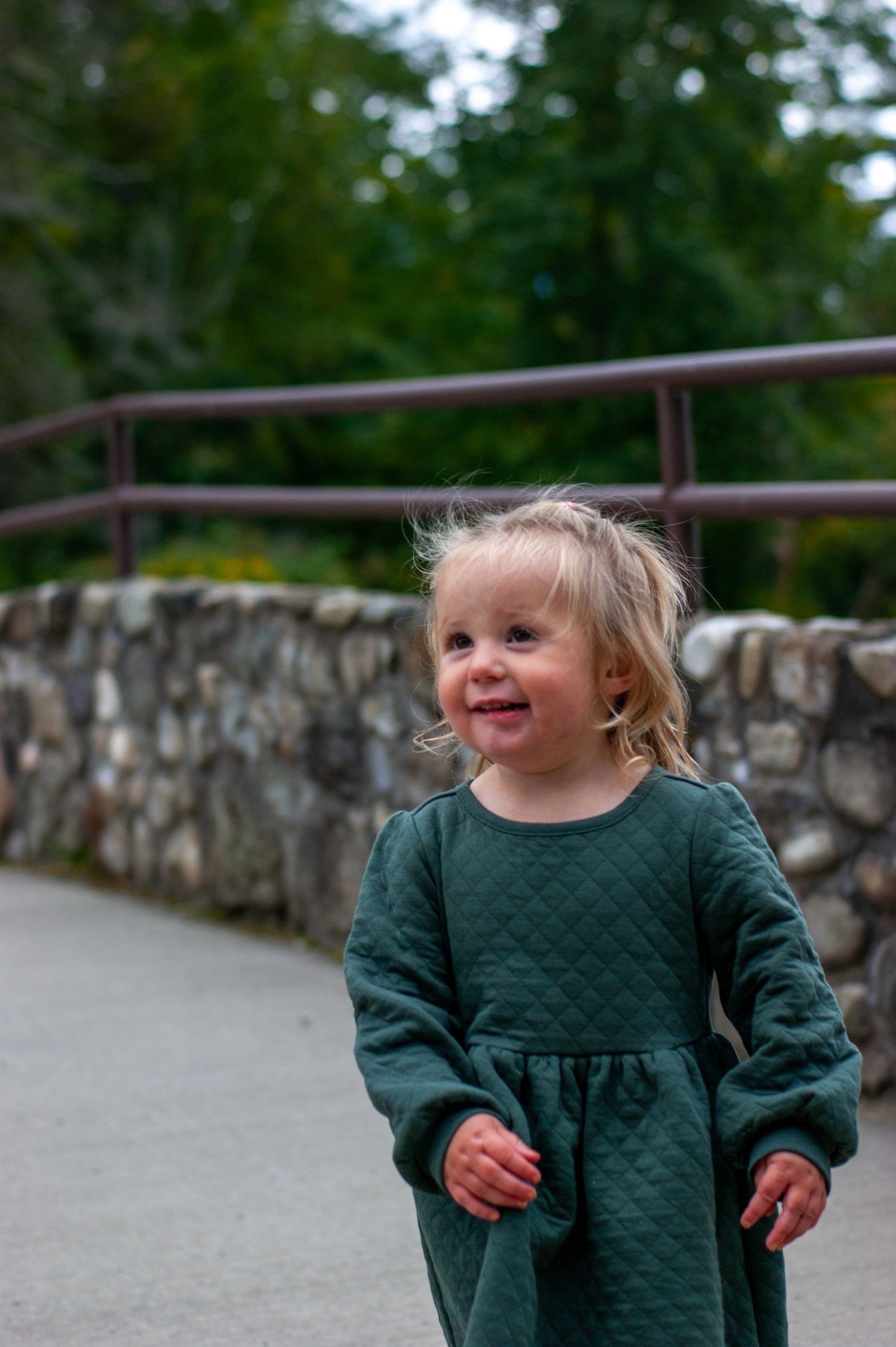 A toddler smiles while standing on a stone bridge path.
