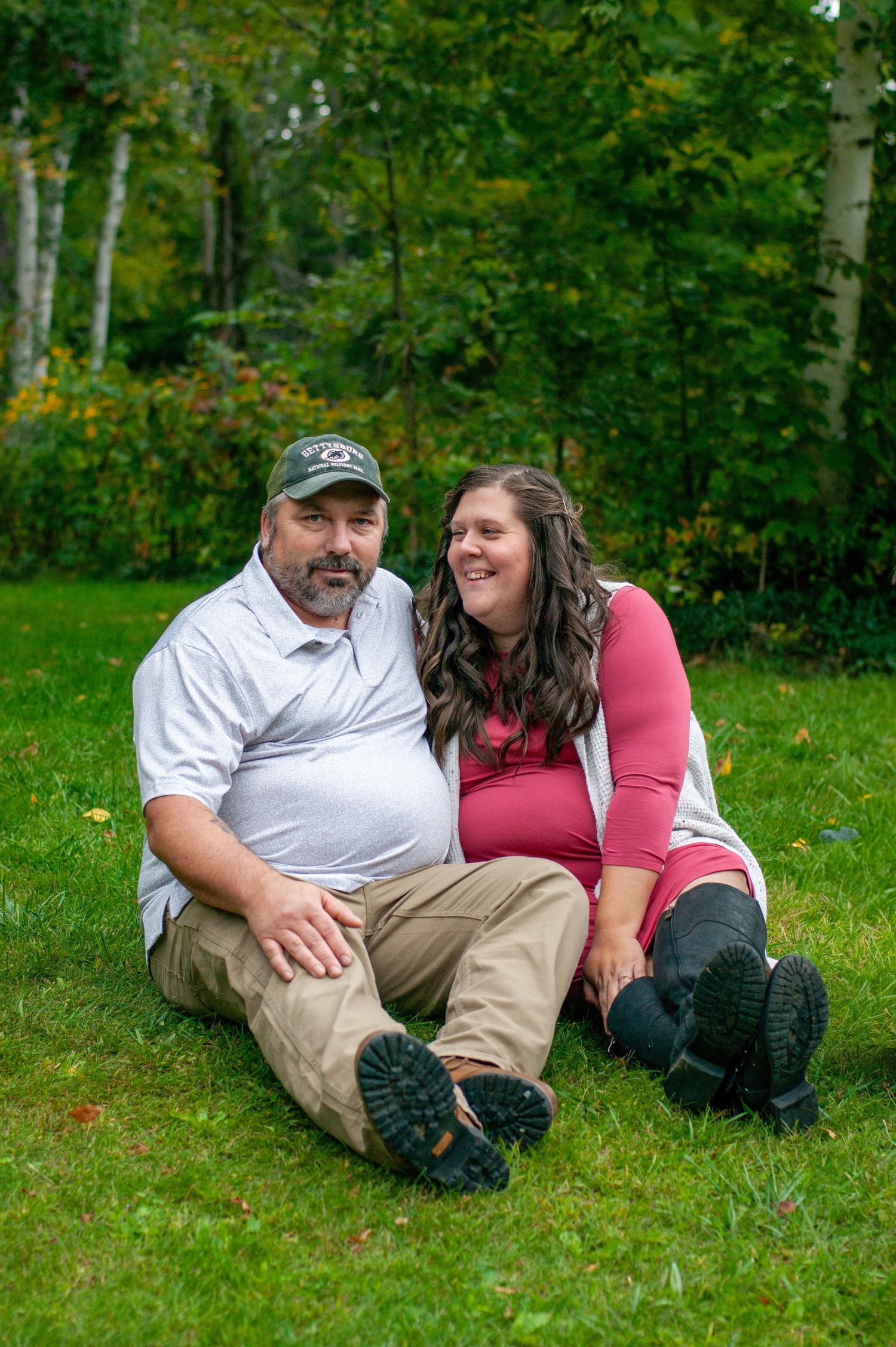 A couple sits together on green grass with autumn trees in the background.