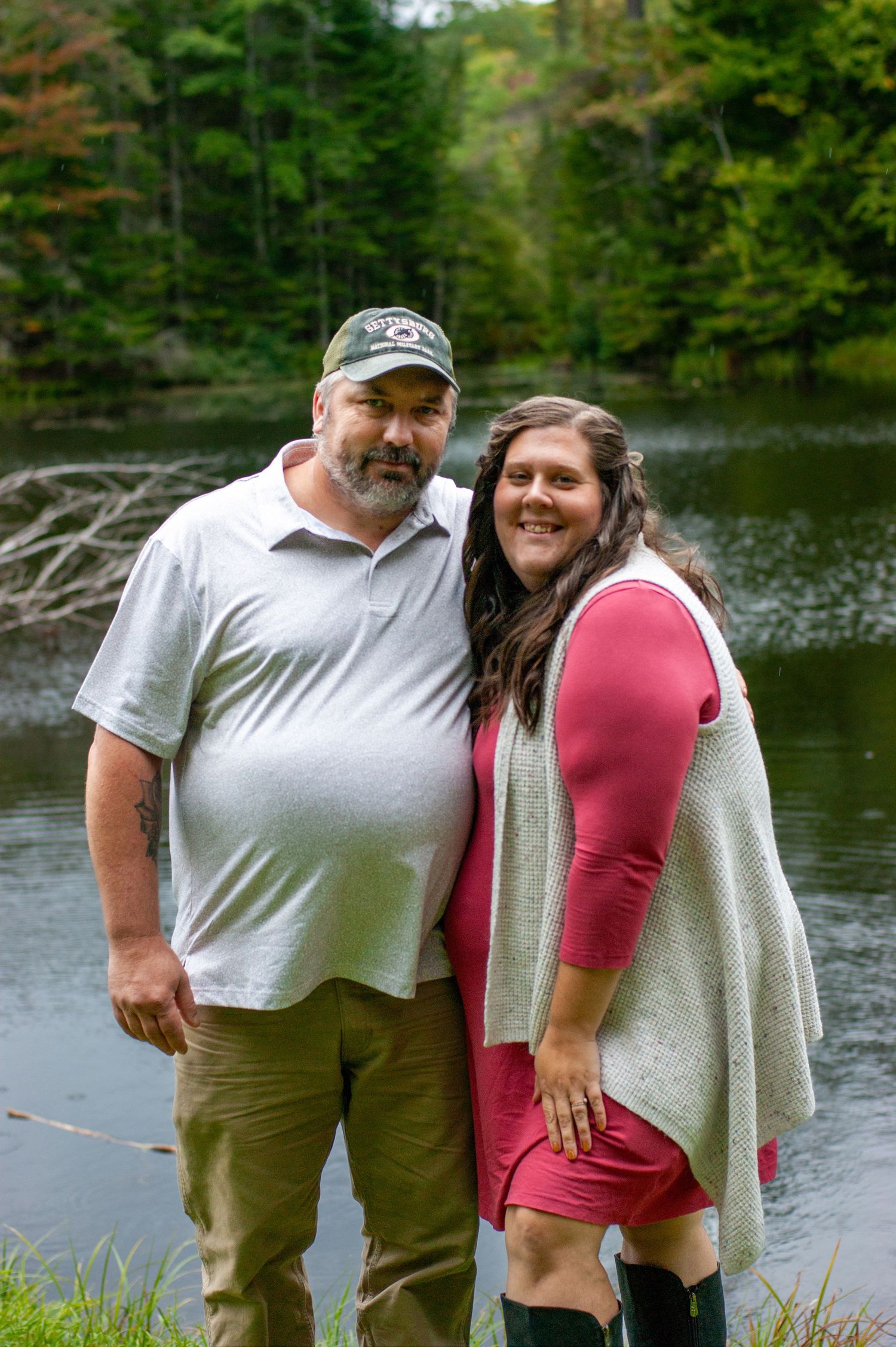 A couple stands by a peaceful lake surrounded by pine trees in autumn.