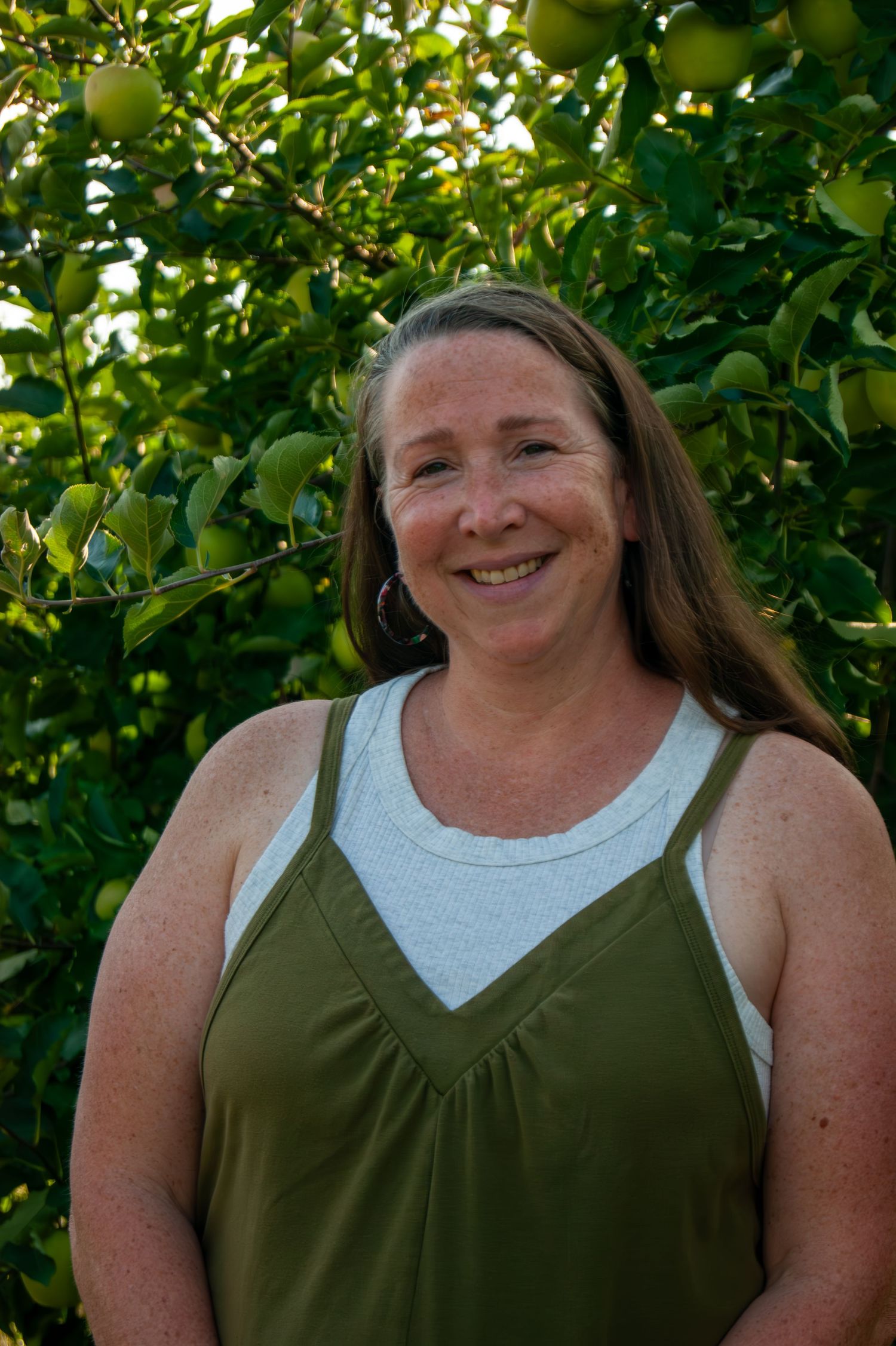 A person in a green tank top stands smiling in front of an apple tree.