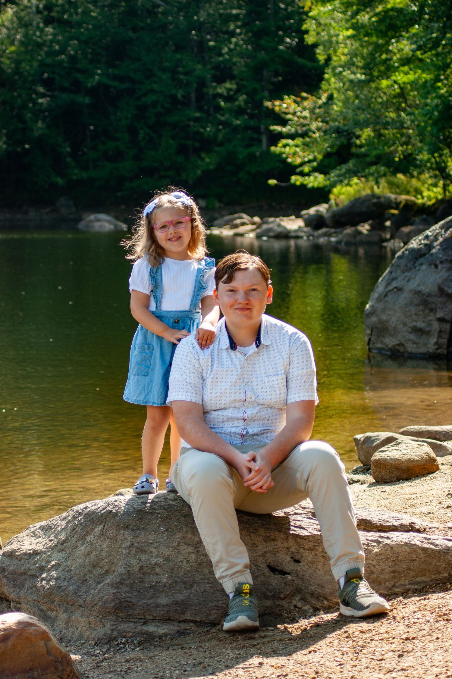 Two people share a heartwarming moment while sitting on rocks by a serene lake surrounded by trees.