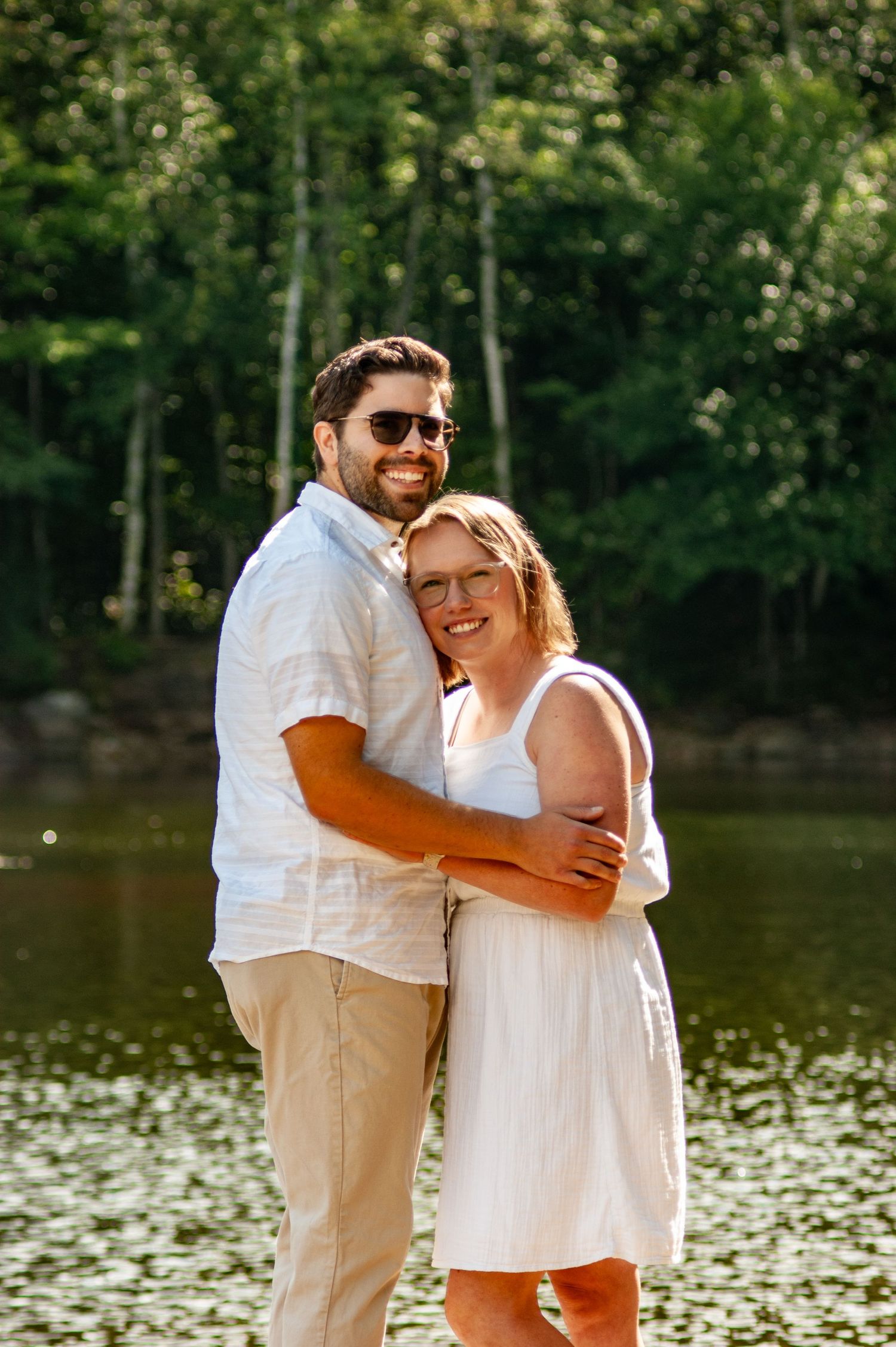 A couple  embraces while standing by a peaceful lake on a sunny summer day.
