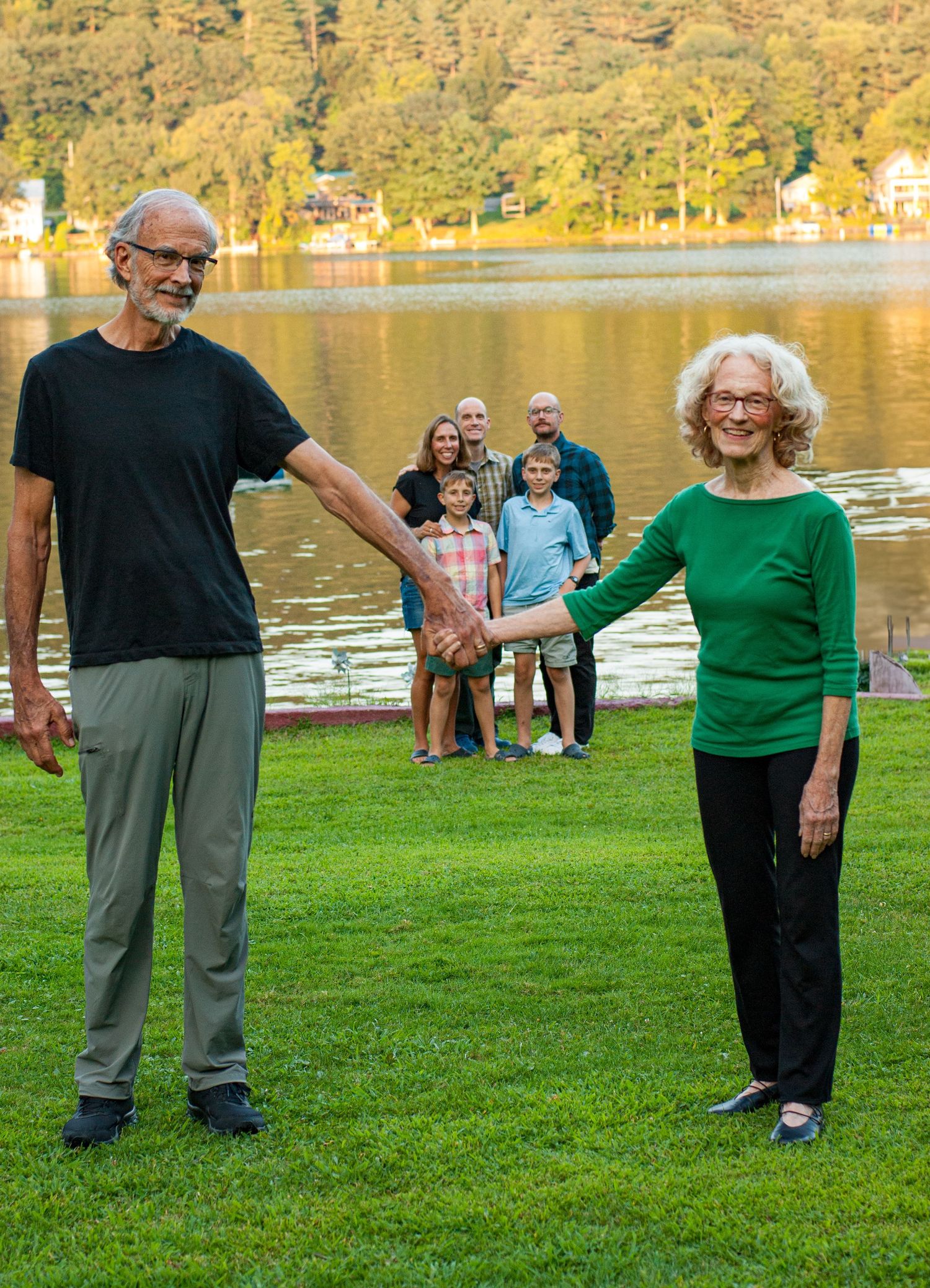 Two people stand on grass near a lake while family members pose in the background during golden hour.