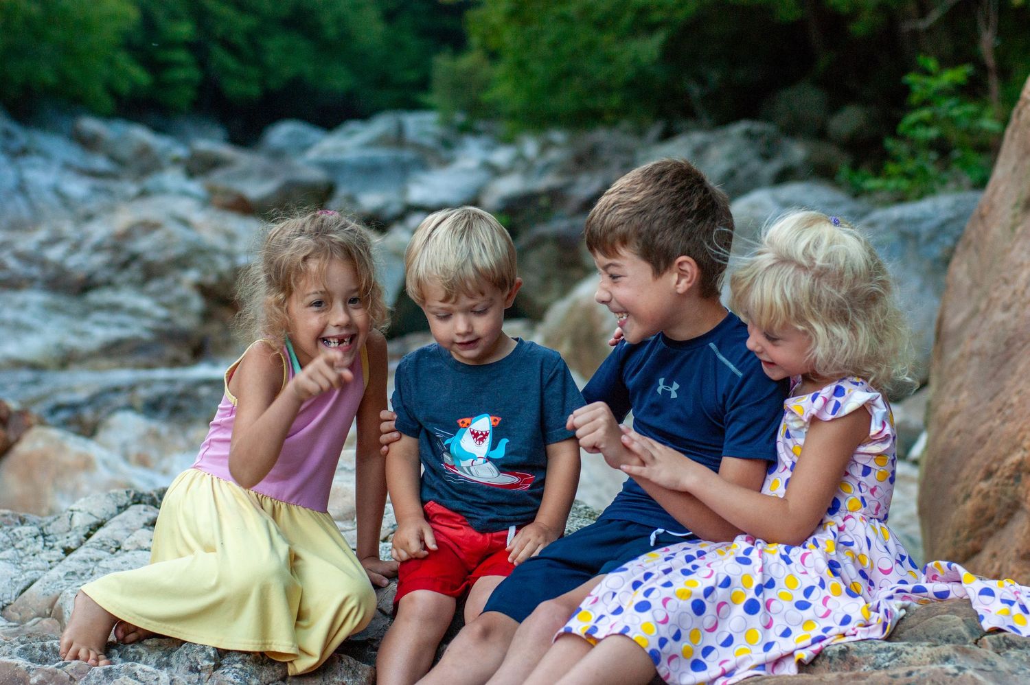 Children sitting on rocks by a stream, enjoying snacks and conversation together.