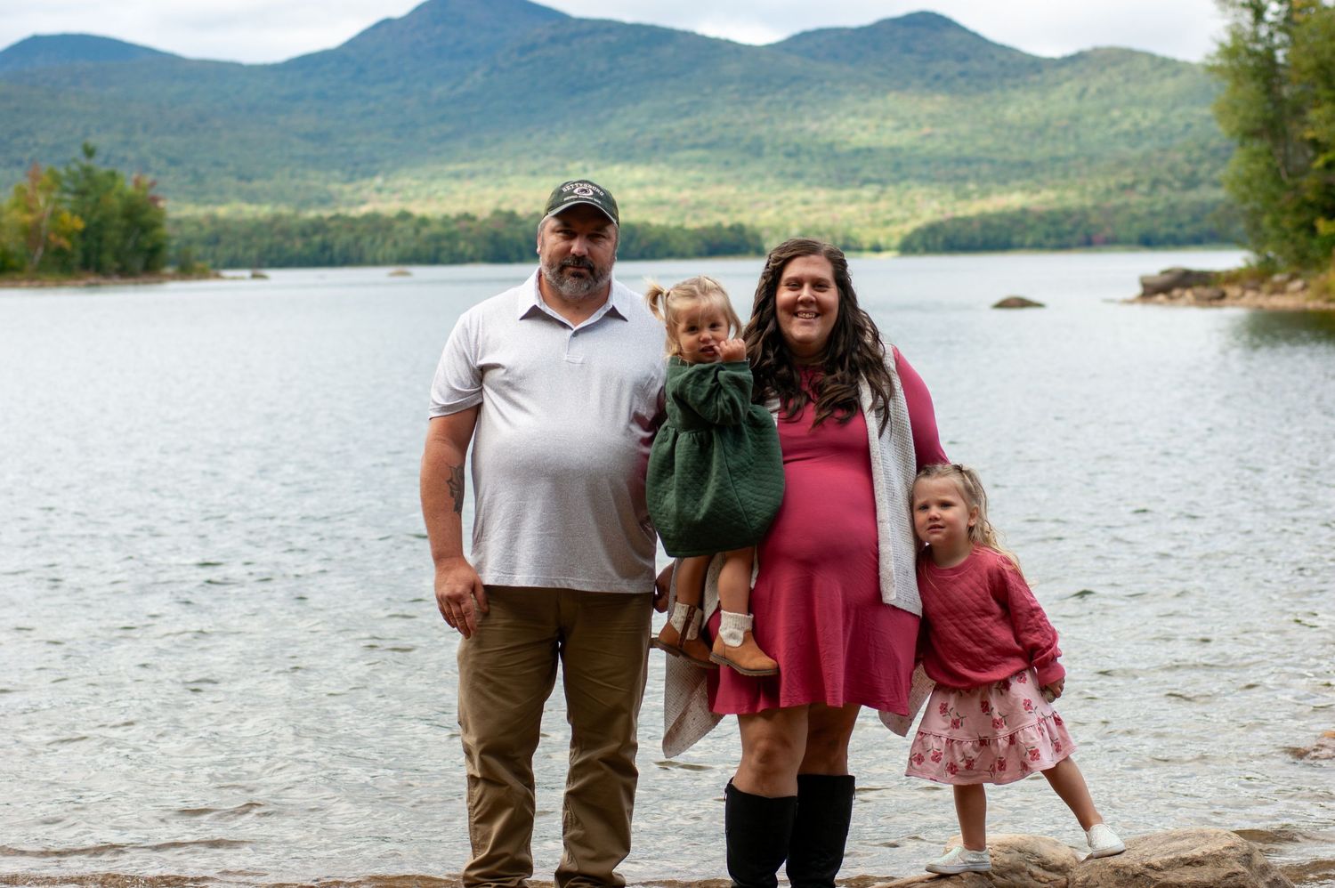 A family poses together by a scenic mountain lake with trees and mountains in the background.