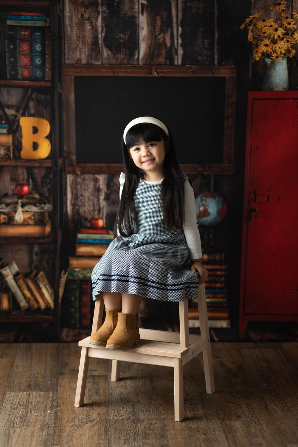 Young person in gray dress and boots sitting on wooden step stool against vintage backdrop.