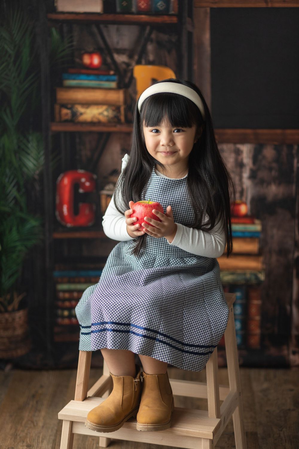 Young child in gray dress and boots sitting on white chair holding red apple in vintage classroom setting.