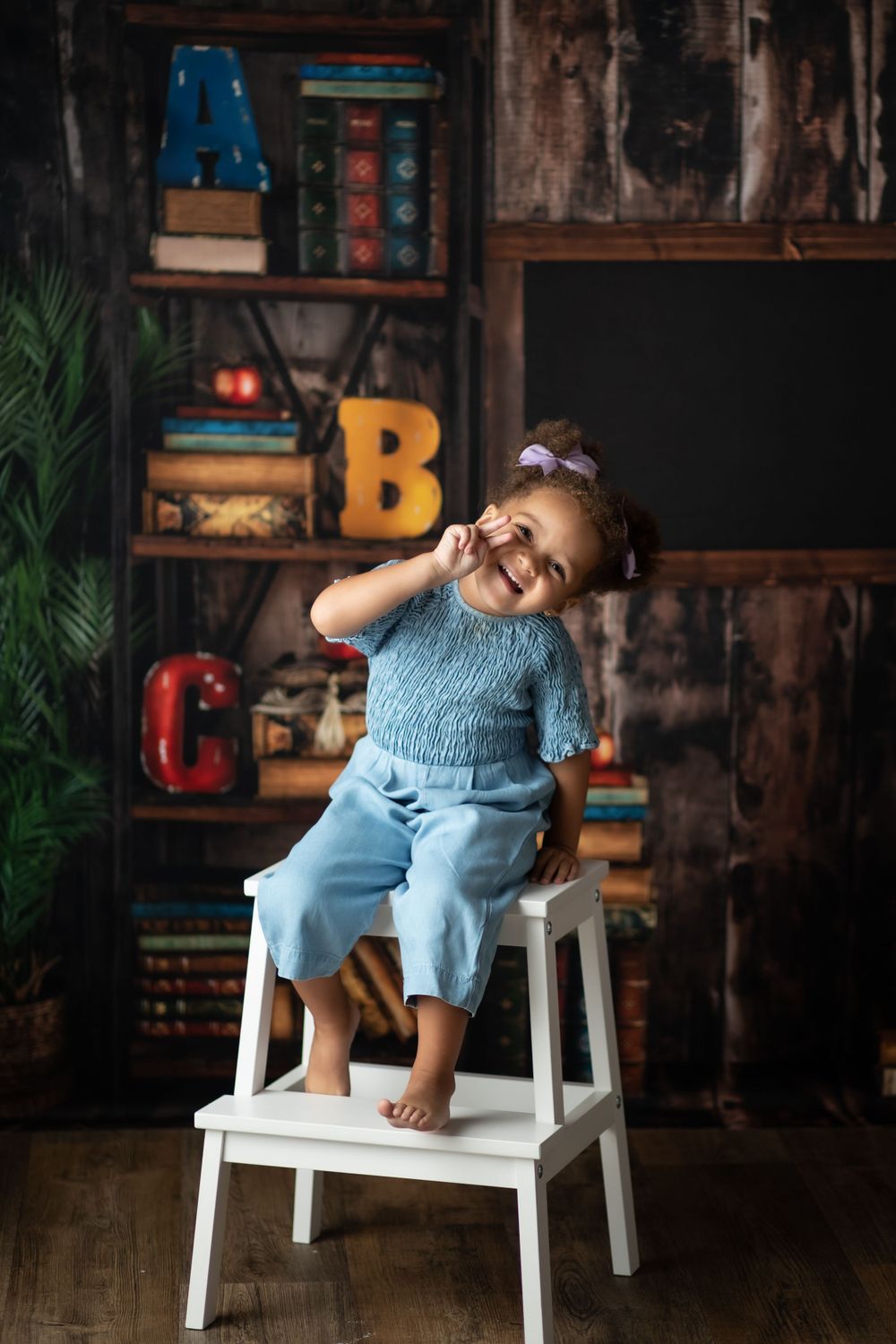 Child in blue outfit sitting on white stepstool with vintage classroom decor and letters in background.