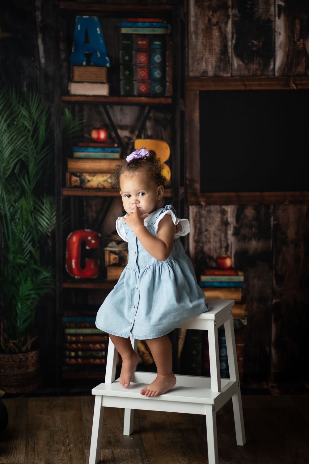 Small child in light blue dress standing on white stepstool against rustic classroom backdrop.