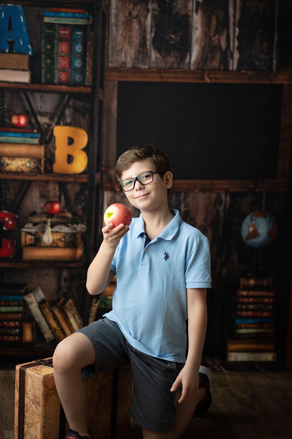 Student in light blue polo and shorts holding apple near vintage bookshelf with decorative letters.