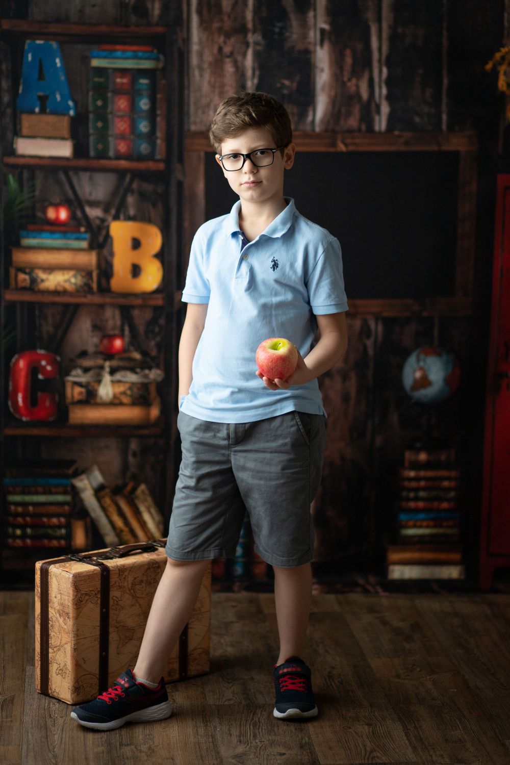 Student in casual attire standing with apple against rustic classroom backdrop with decorative letters.
