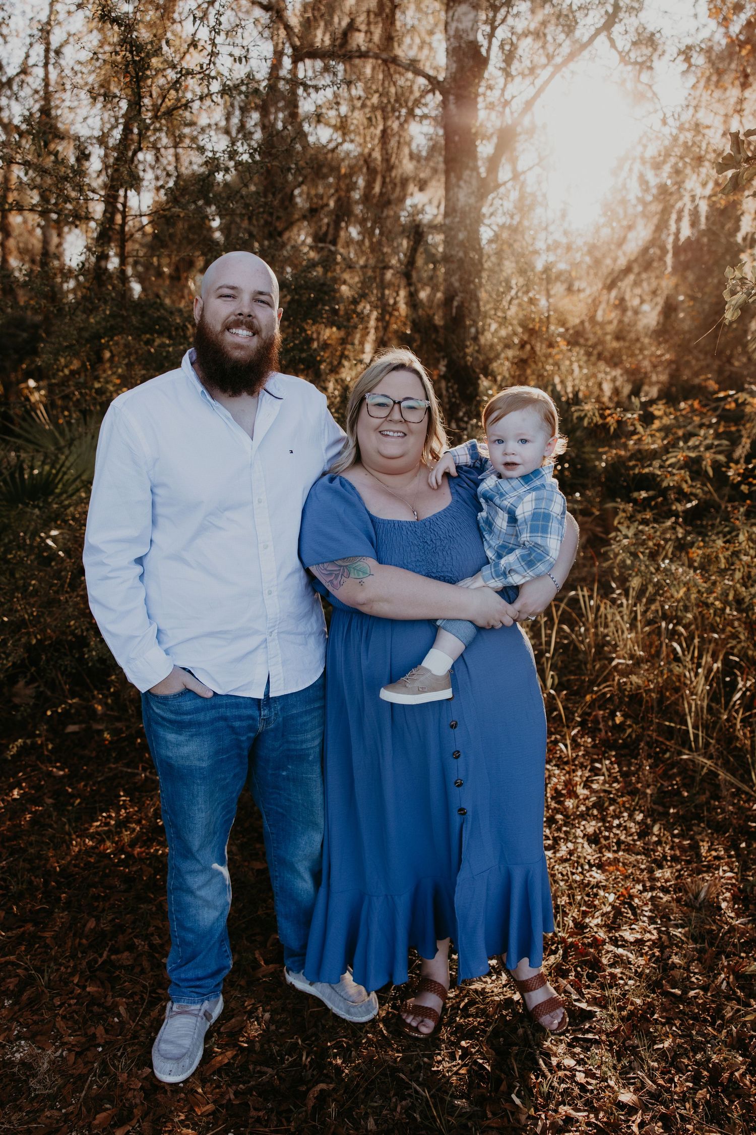 Intimate maternity photo of couple in flowing blue dress amid pine forest at sunset.